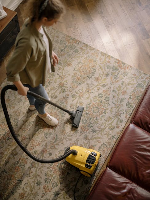 Close-up of a vacuum cleaner nozzle with a black plastic body, operated by a person wearing dark pants and white shoes, cleaning a patterned carpet in a residential room. The carpet features a floral 